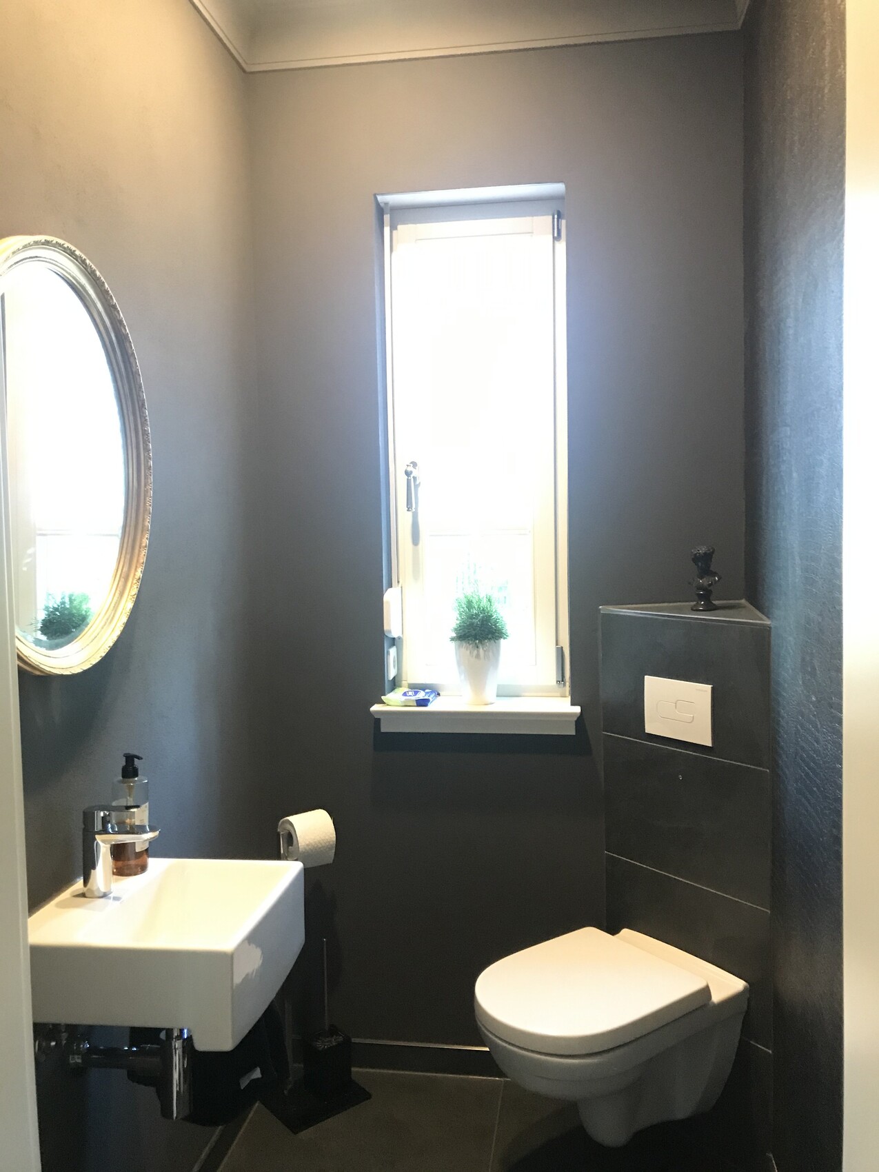 Kleines, modernes Badezimmer mit dunklen Wänden, WC, Waschbecken, Spiegel und Pflanze am Fenster.