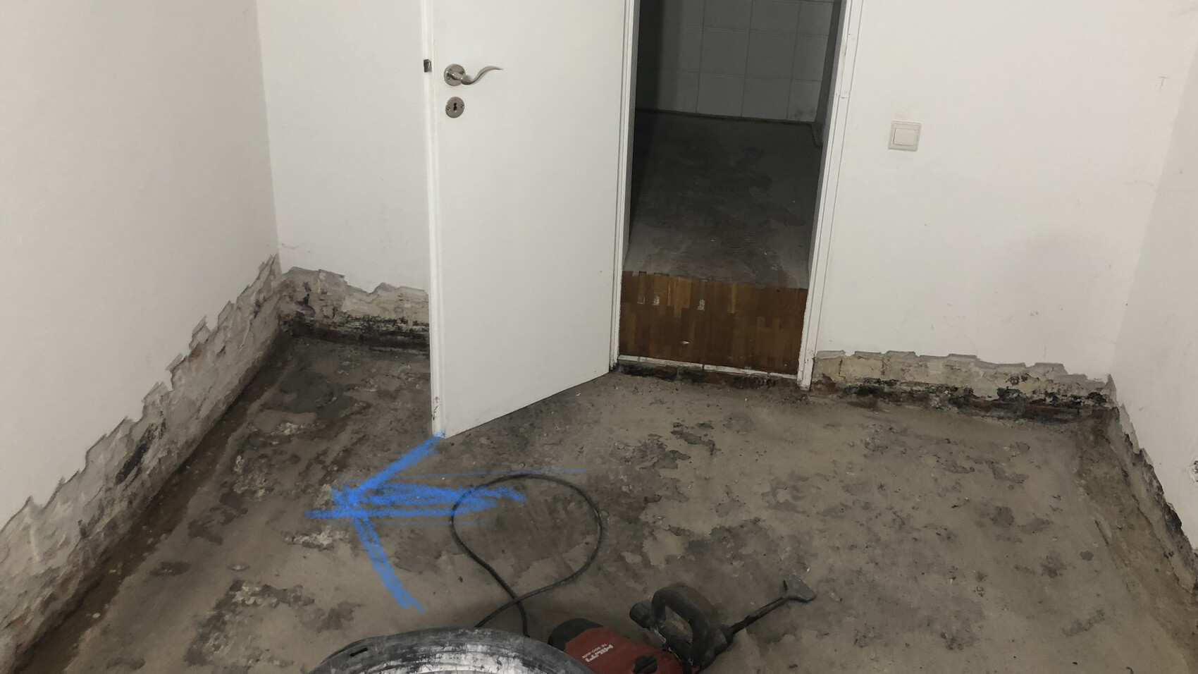 Room with damaged floor, exposed plaster at the base; door to a dark room, blue arrow