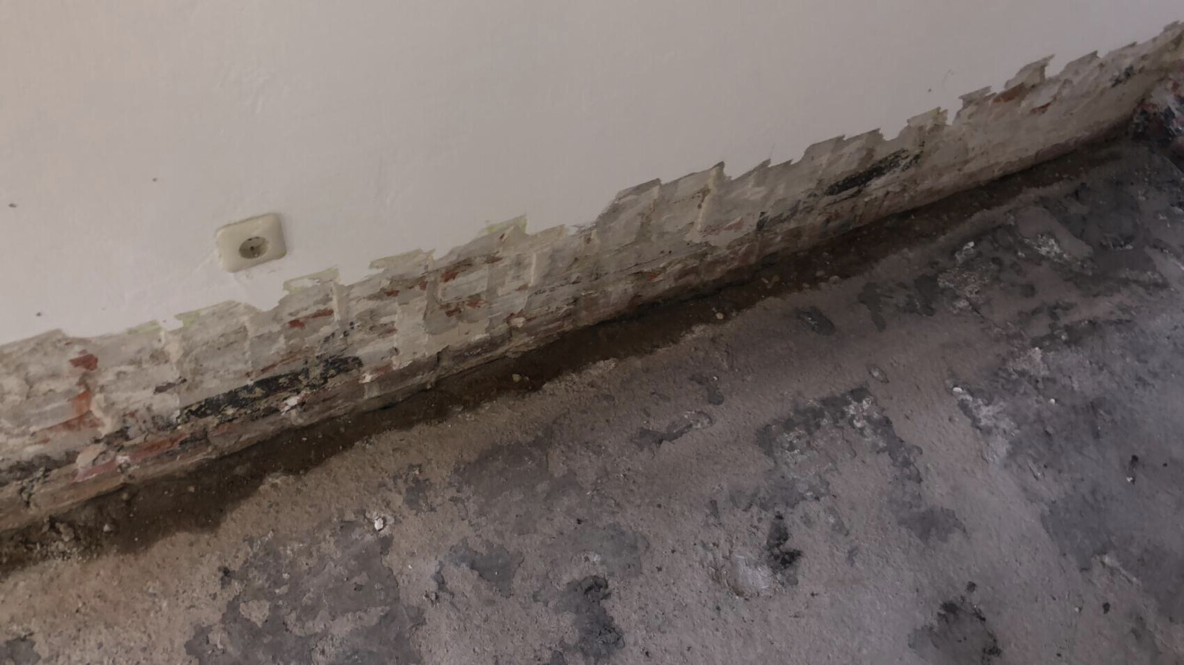 Broken plaster at the lower wall area; exposed bricks underneath, damp floor and electrical outlet.