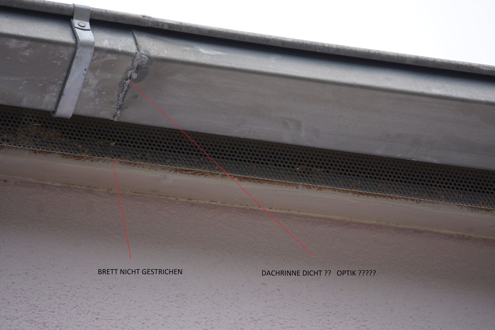 Close-up of the eaves with metal gutter, perforated metal ventilation, and damaged board cladding.
