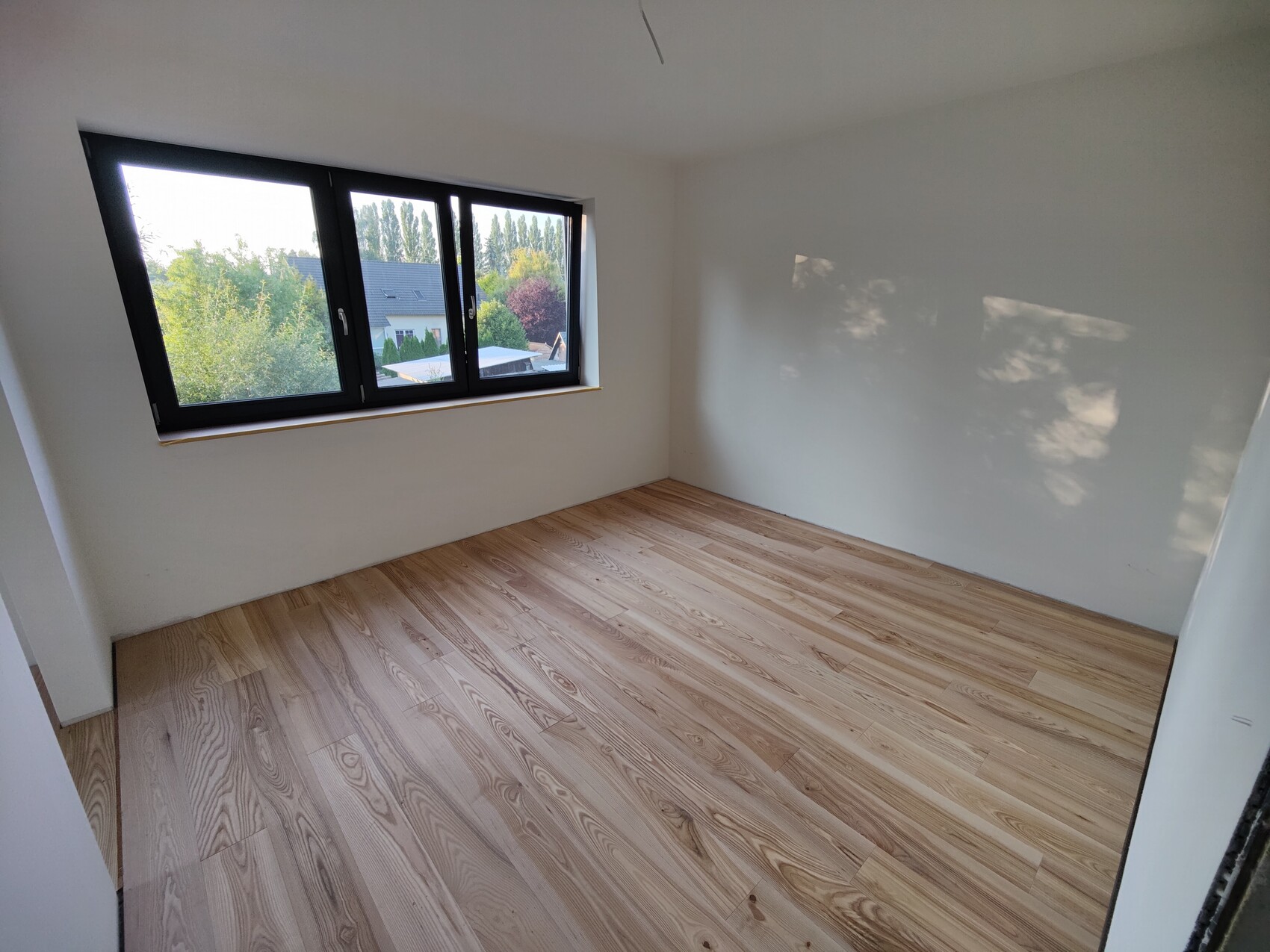 Leeres Zimmer mit hellen Wänden, HolzfuĂboden und groĂem dreiteiligem Fenster mit Blick auf Bäume.