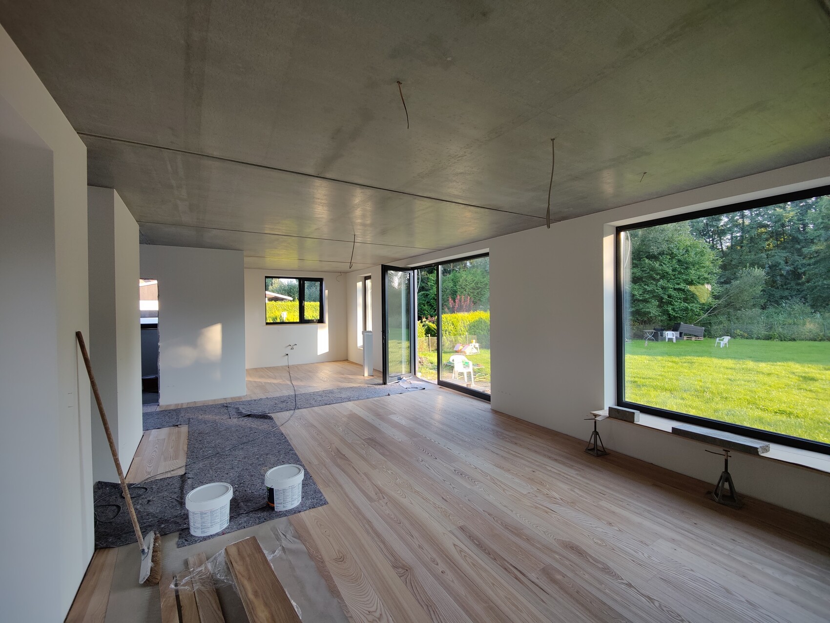 GroĂer Bauinnenraum mit roher Decke, HolzfuĂboden, Eimern, Besen; Blick auf Garten durch Fenster.