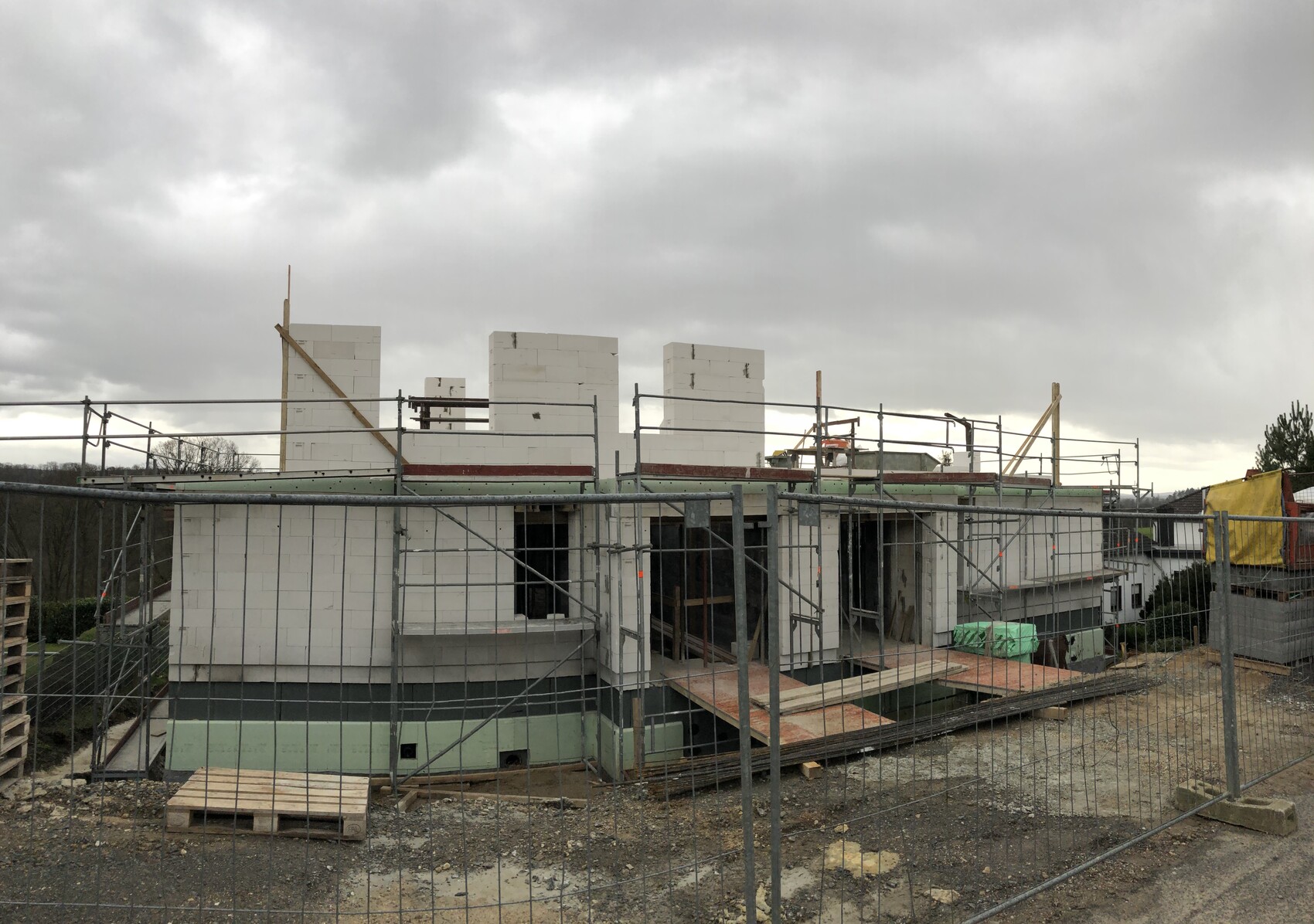 Baustelle mit weißen Blockwänden, Gerüst, Holzplanken und Paletten unter grauem Himmel.