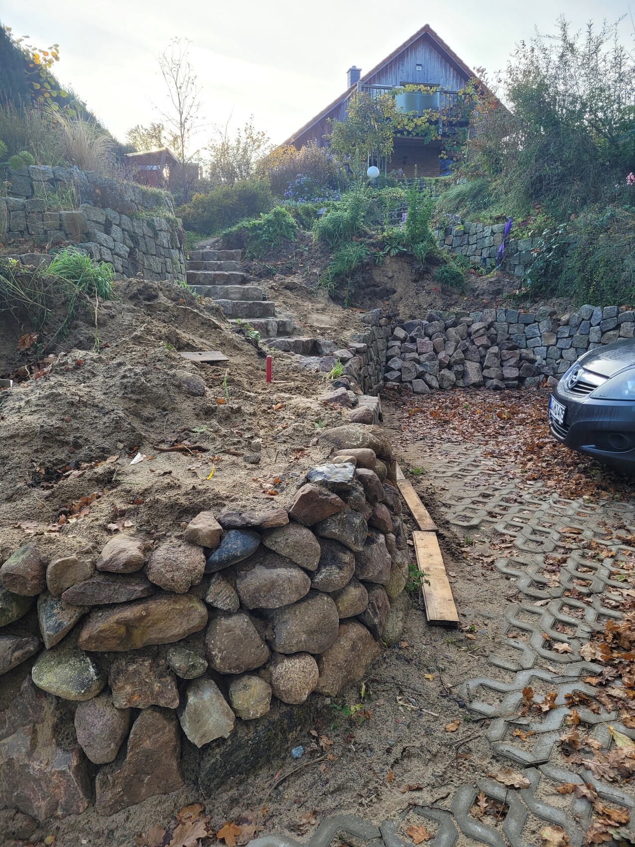 Garten mit Steinmauern, Stufen zum Holzhaus oben; rechts geparktes Auto.