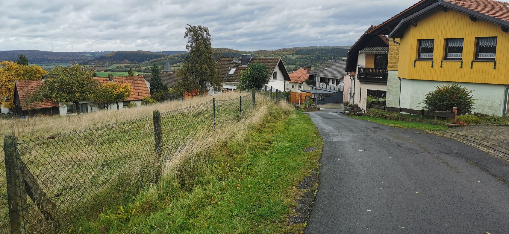 Rural village street with yellow house on the right, fence on the left, grassland and wind turbines in the background.