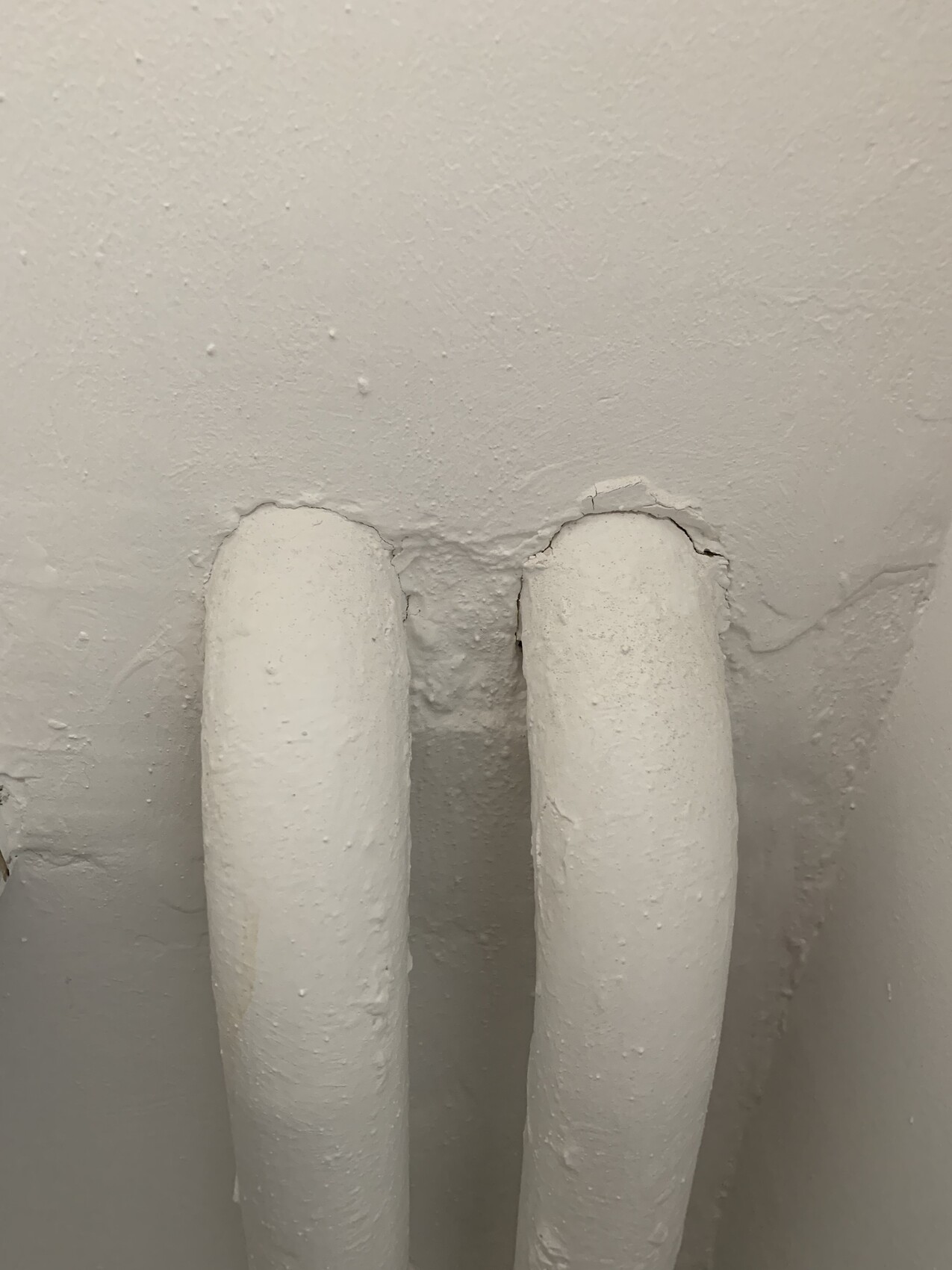 Two white pipes protrude from a rough wall; plaster is broken around the pipes.