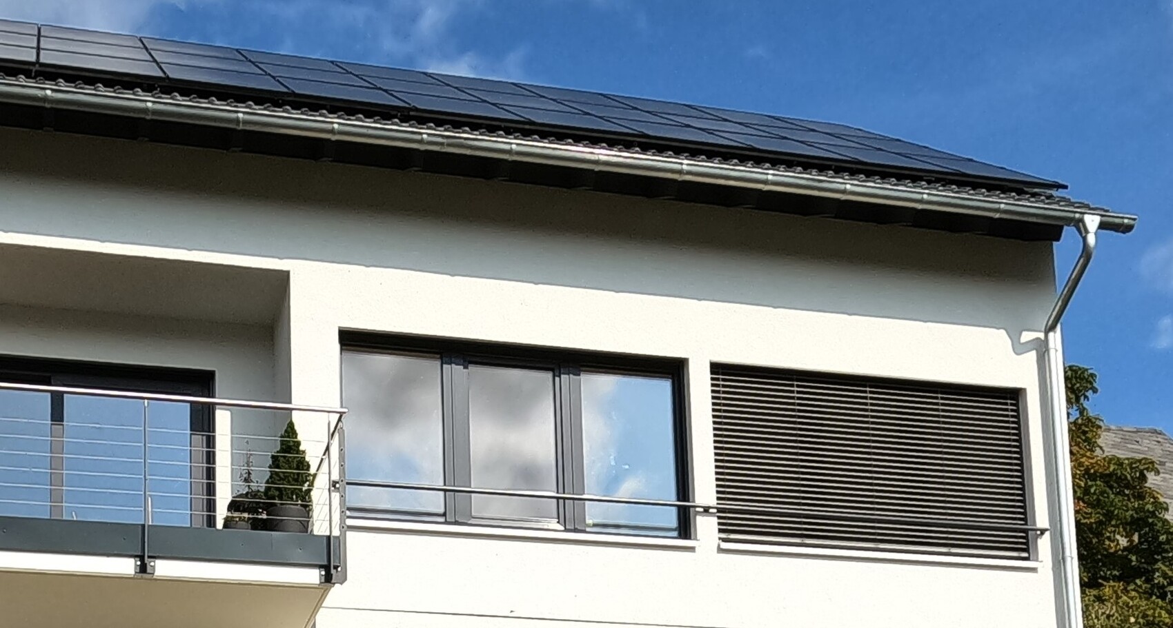 Modern house facade with balcony, large windows, roller shutters, and solar panels on the roof