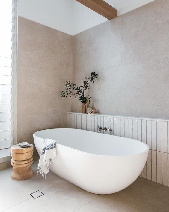 Freestanding white bathtub in a bright bathroom; green plant, towel and wooden stool.