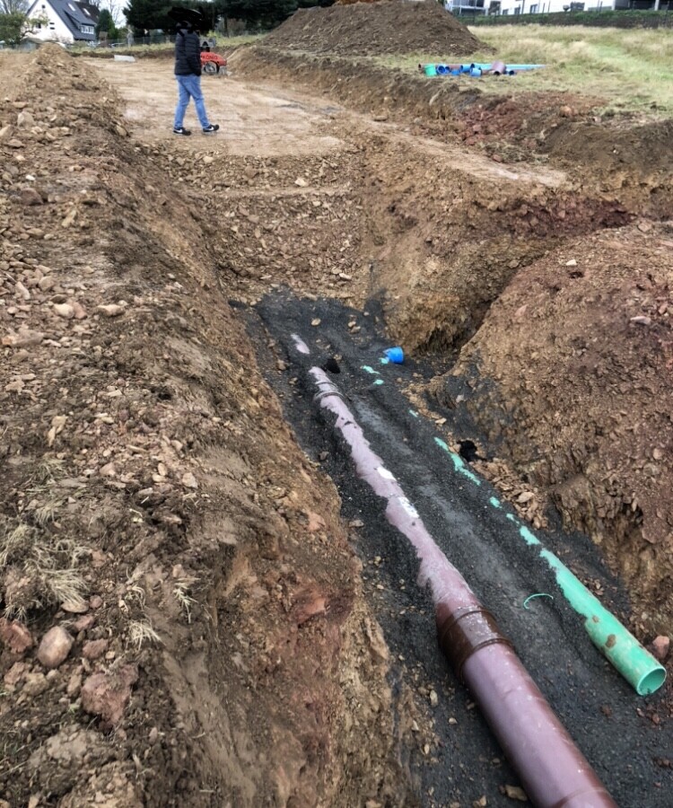 Construction site with excavated trenches, pipes, and large soil piles; workers in the background.