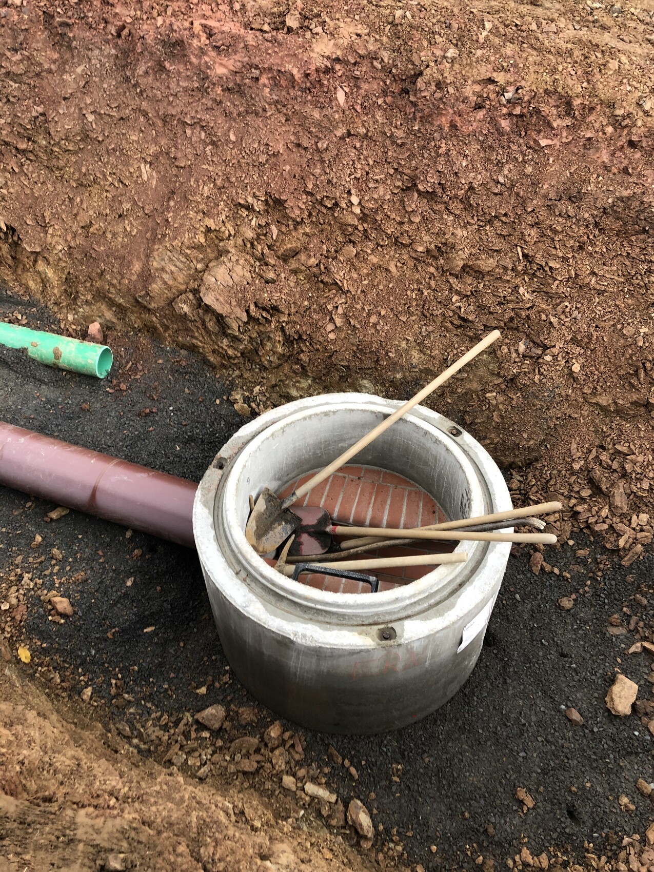 Round concrete channel cover in a trench with brick base, wooden sticks and pipes nearby.