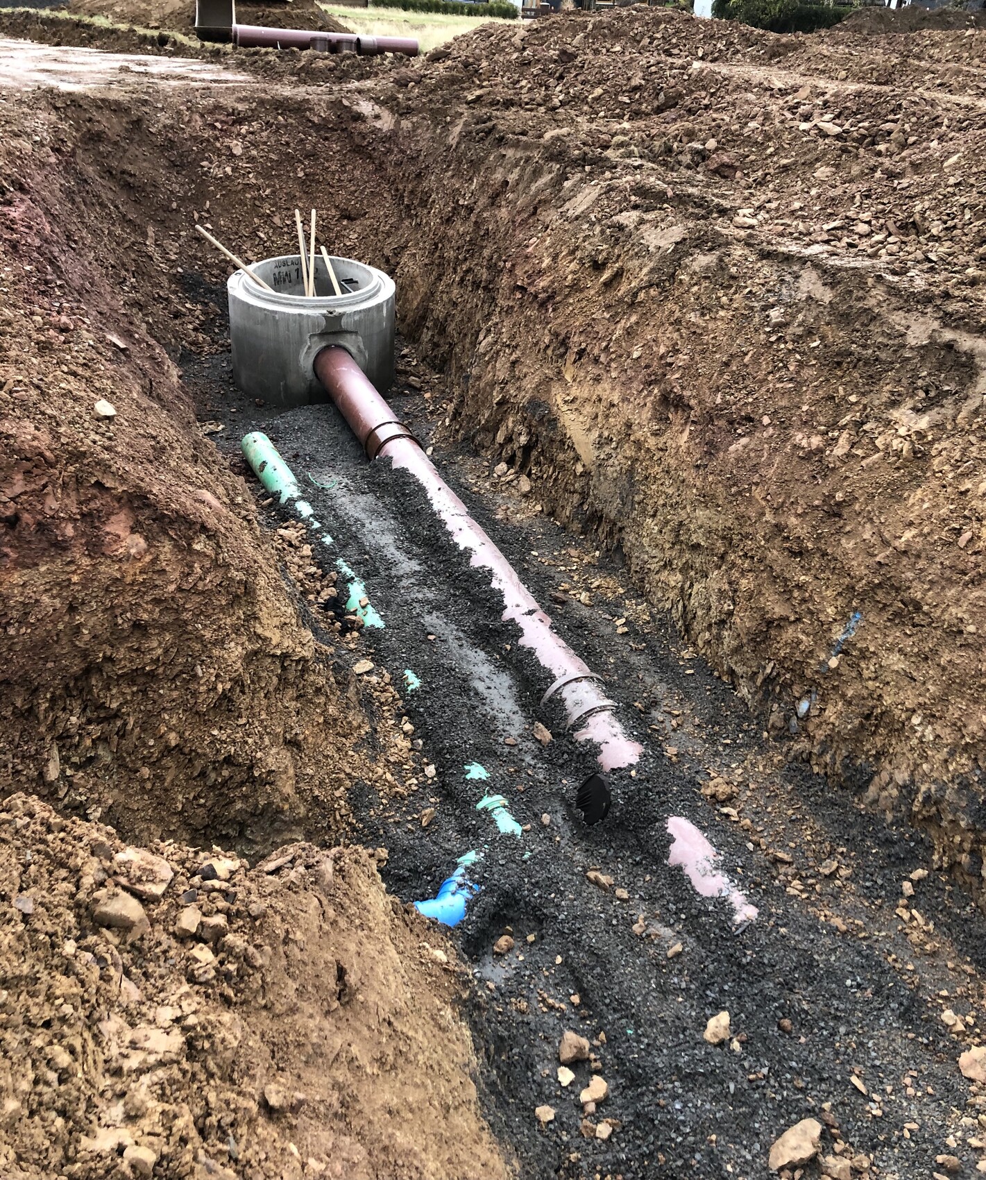 Excavation pit with pink PVC pipe, round concrete channel and wooden stakes in gravel.