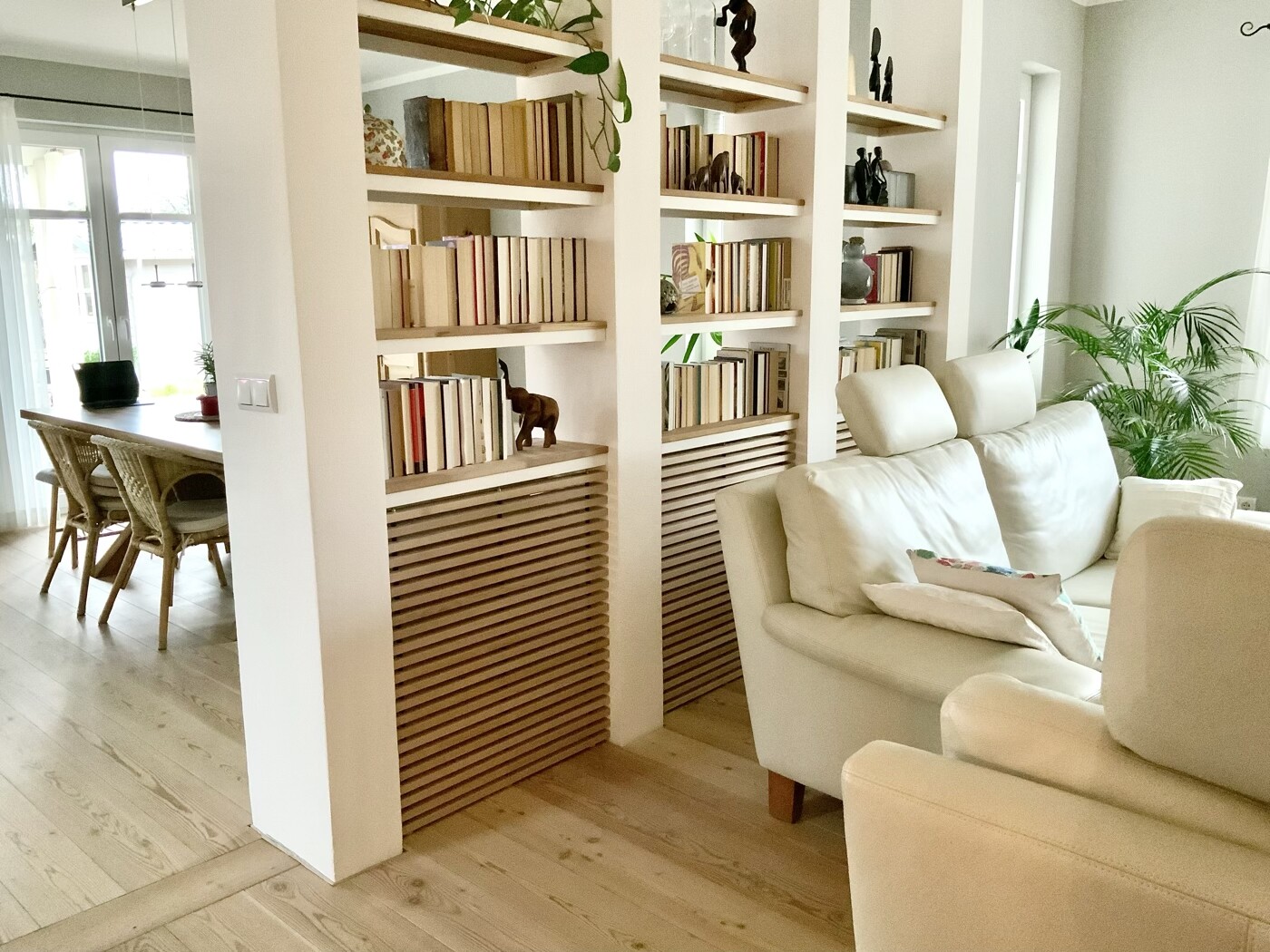 Heller Wohnraum mit weißem Sofa, offenen Bücherregalen und großen Grünpflanzen.