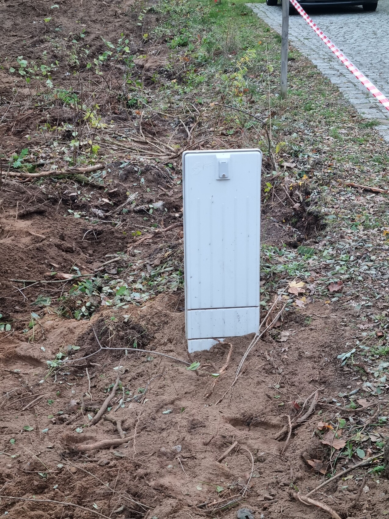 White metal distribution box standing in an open excavation pit, barrier tape in the background.
