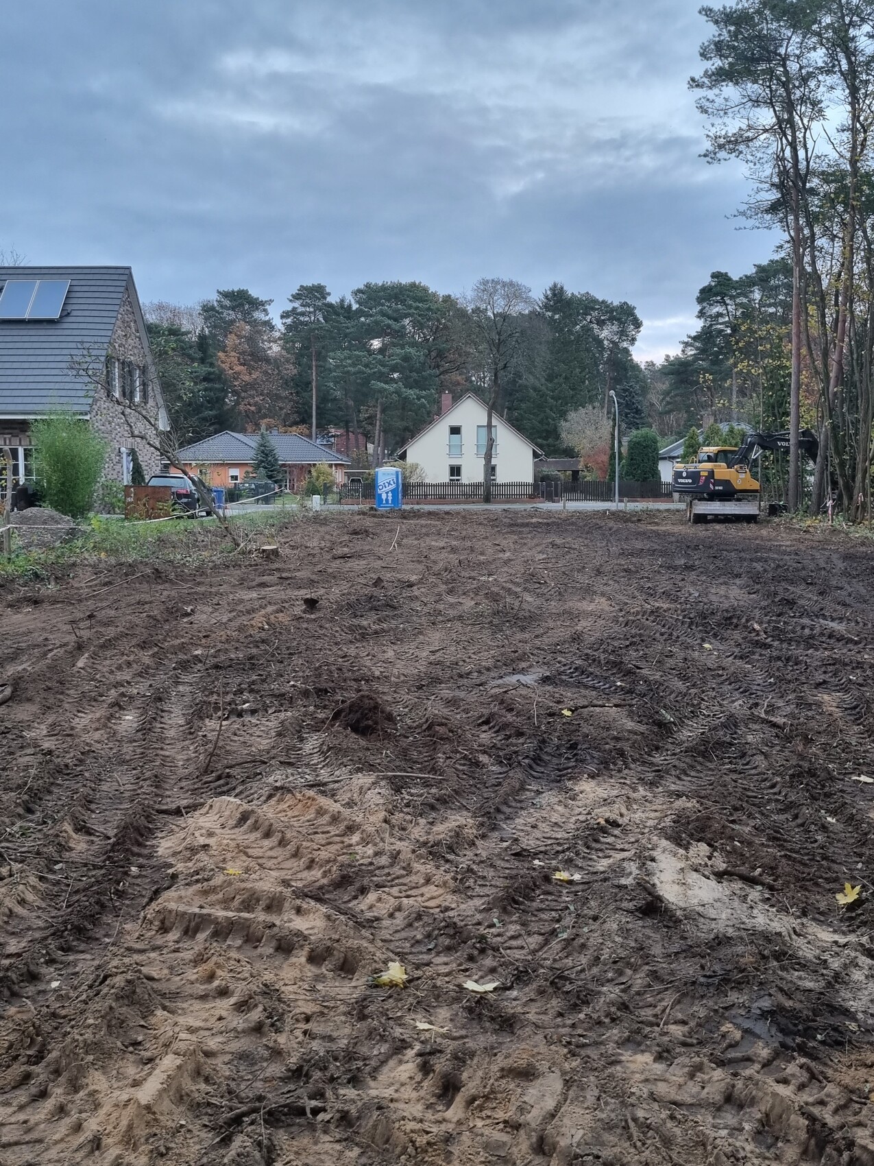 Construction site with excavated soil, excavator on the right, residential houses and forest in the background.