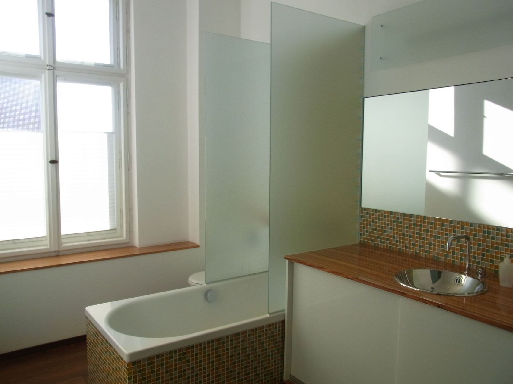 Bright bathroom with bathtub, glass shower screen, wooden countertop and mosaic tiles.
