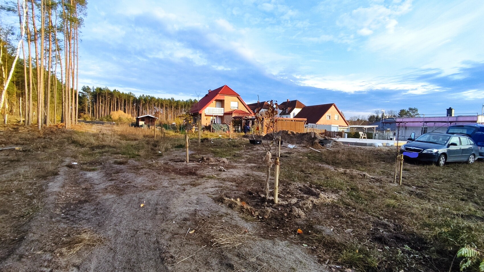 Building area with red tiled roofs, construction work, row of trees on the left, parked cars on the right.