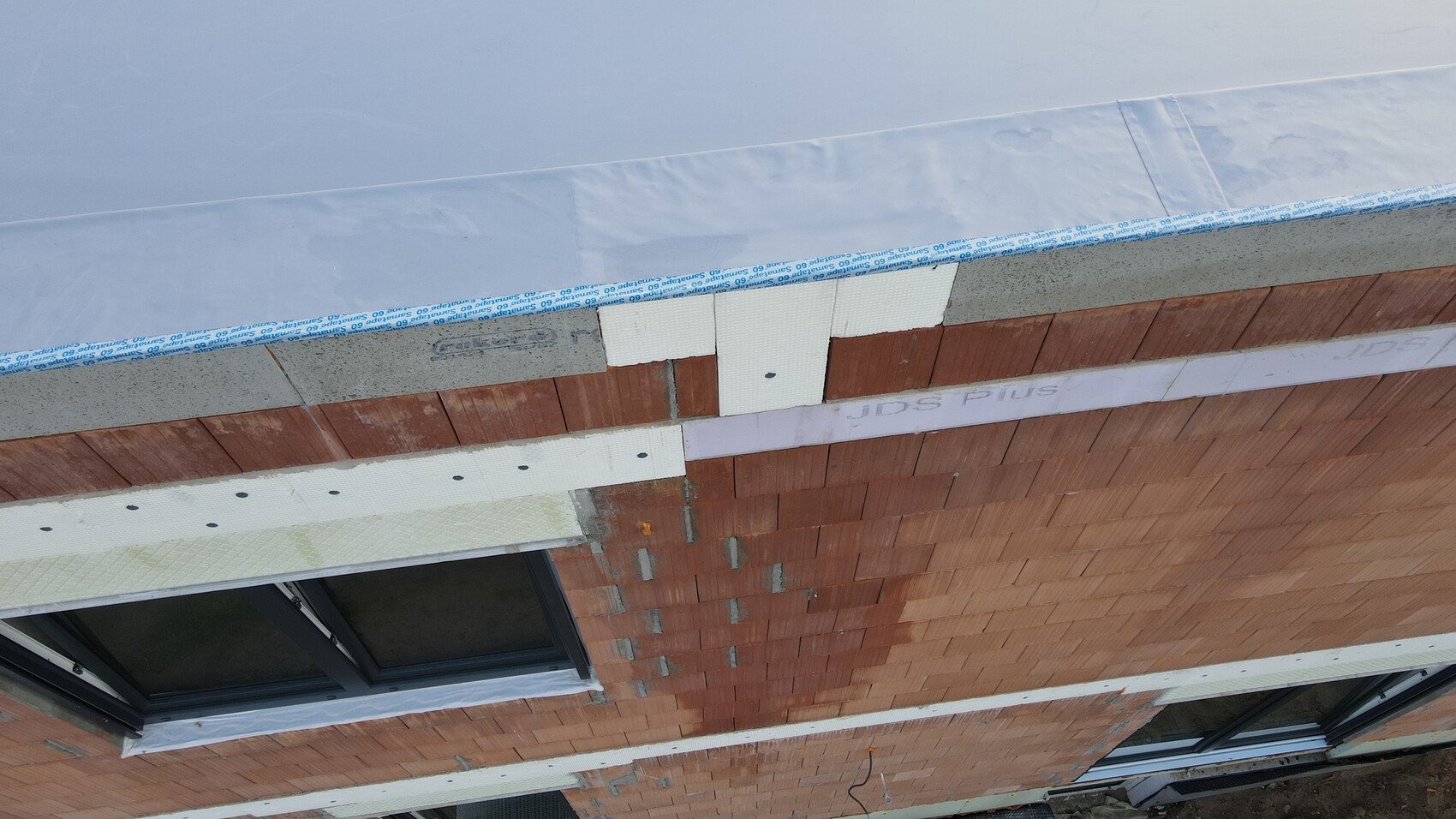 Außenwand-Baustelle: rote Ziegel, Dämmplatten, weiße Platten, Blaufolie, Fenster unten links