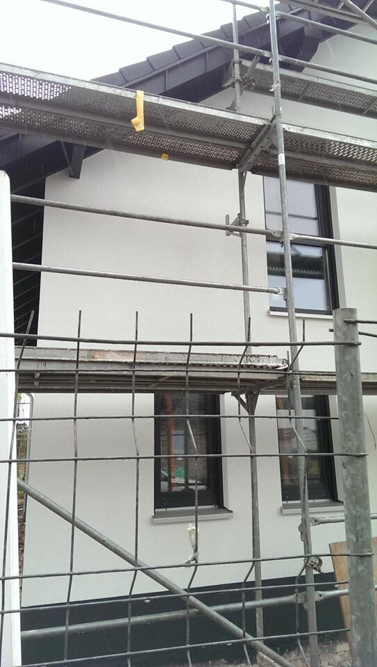 Construction scaffolding in front of a modern white house with windows; construction work visible.