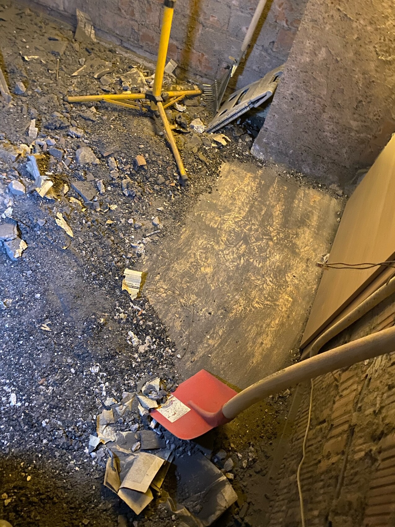 Basement construction site: debris, dust, yellow tripod, red shovel, brick wall.