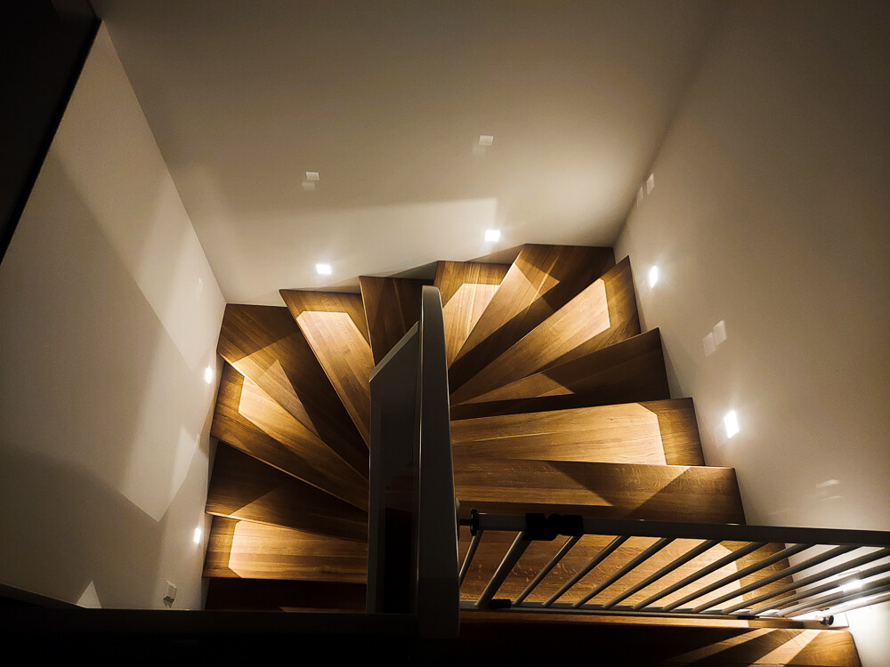 Spiral wooden staircase viewed from above, warmly lit, white walls, railing at the edge.