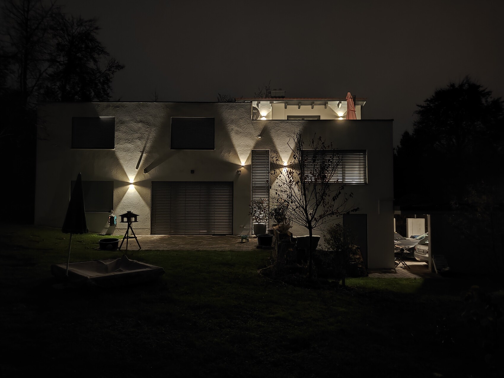 Modern white house at night with outdoor lighting, terrace, and garden in the foreground.