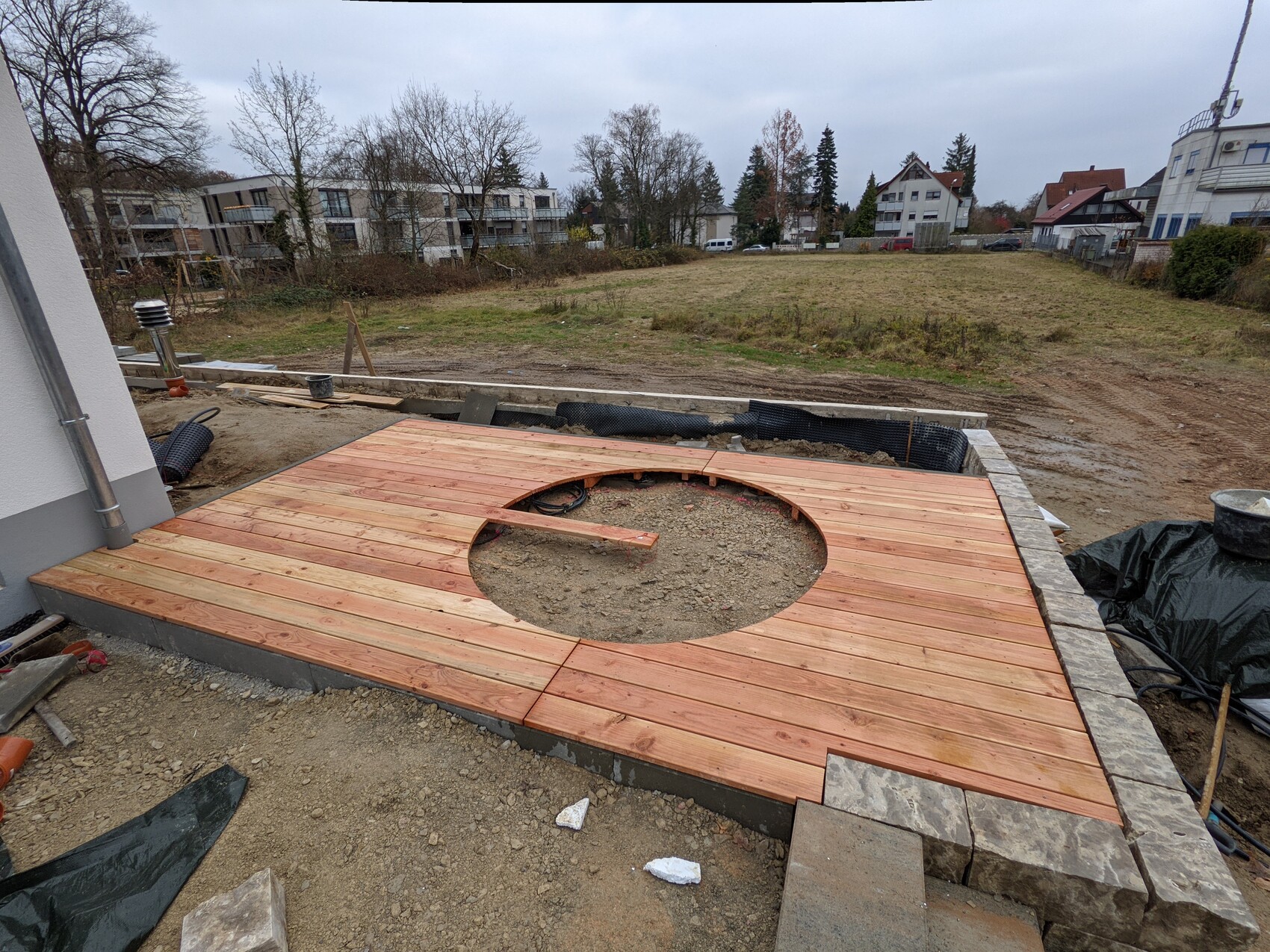 Wooden deck with circular cutout in front of the house; construction site and garden in the background.
