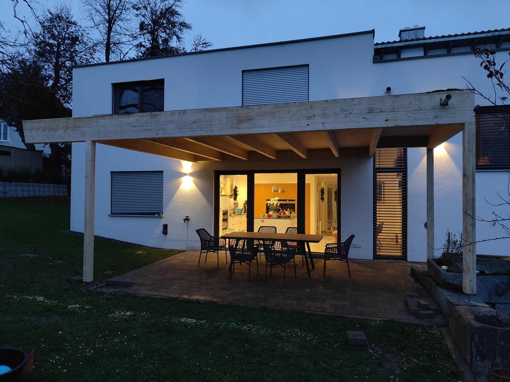 Modern white rear house facade with wooden pergola, outdoor dining table, illuminated interior.