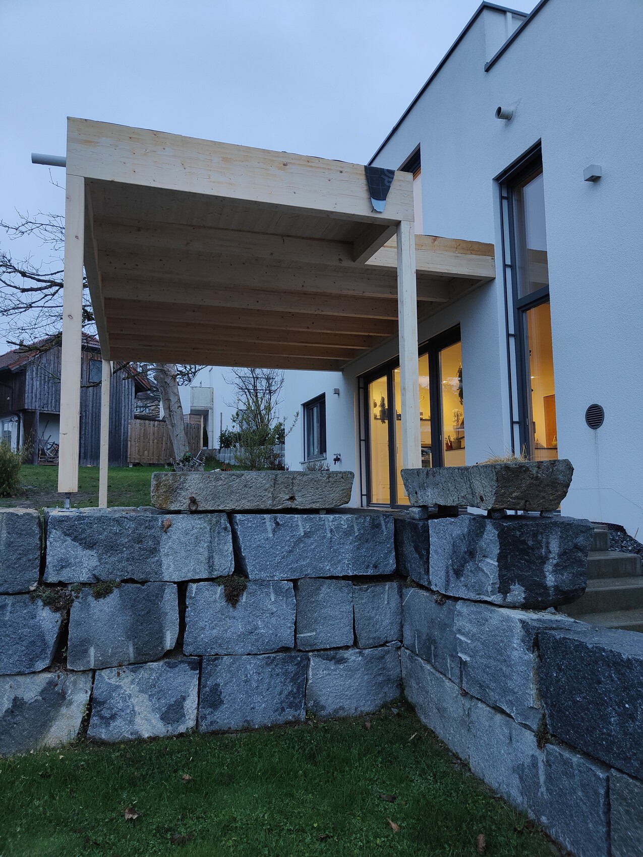 Modern house extension with wooden terrace roof over stone block wall and illuminated interiors.