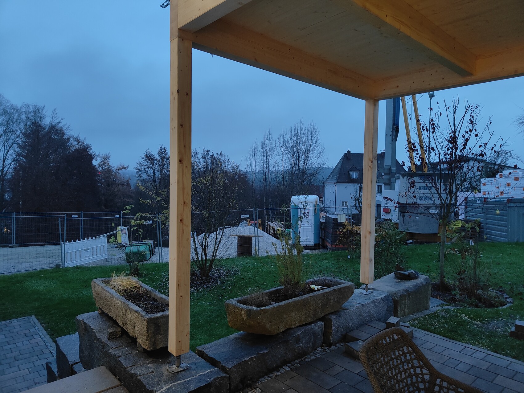 View of terrace with wooden posts and stone planter boxes; construction fence, toilet, and building site in the background.
