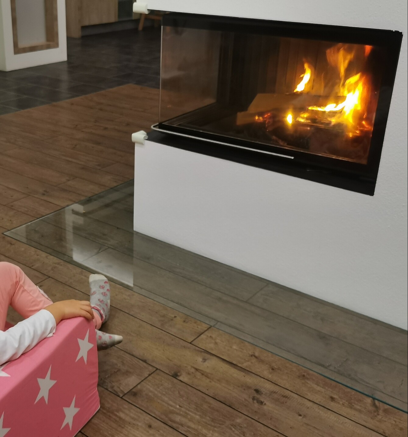 Modern fireplace in white wall with glass front and burning fire; child on pink star-patterned chair.