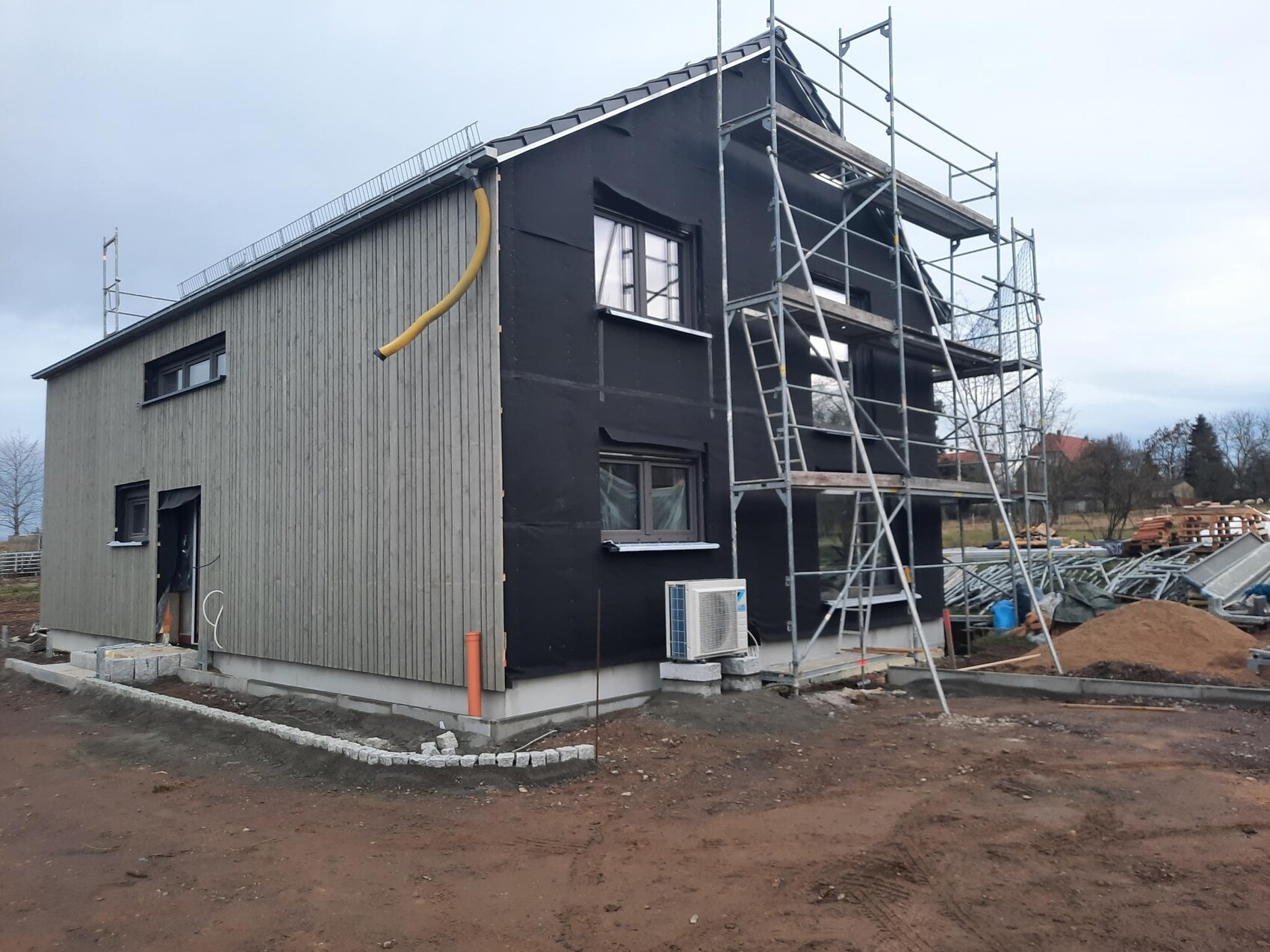 Zweistöckiges Haus im Bau mit Gerüst; linke Holzverkleidung, rechte schwarze Fassade.