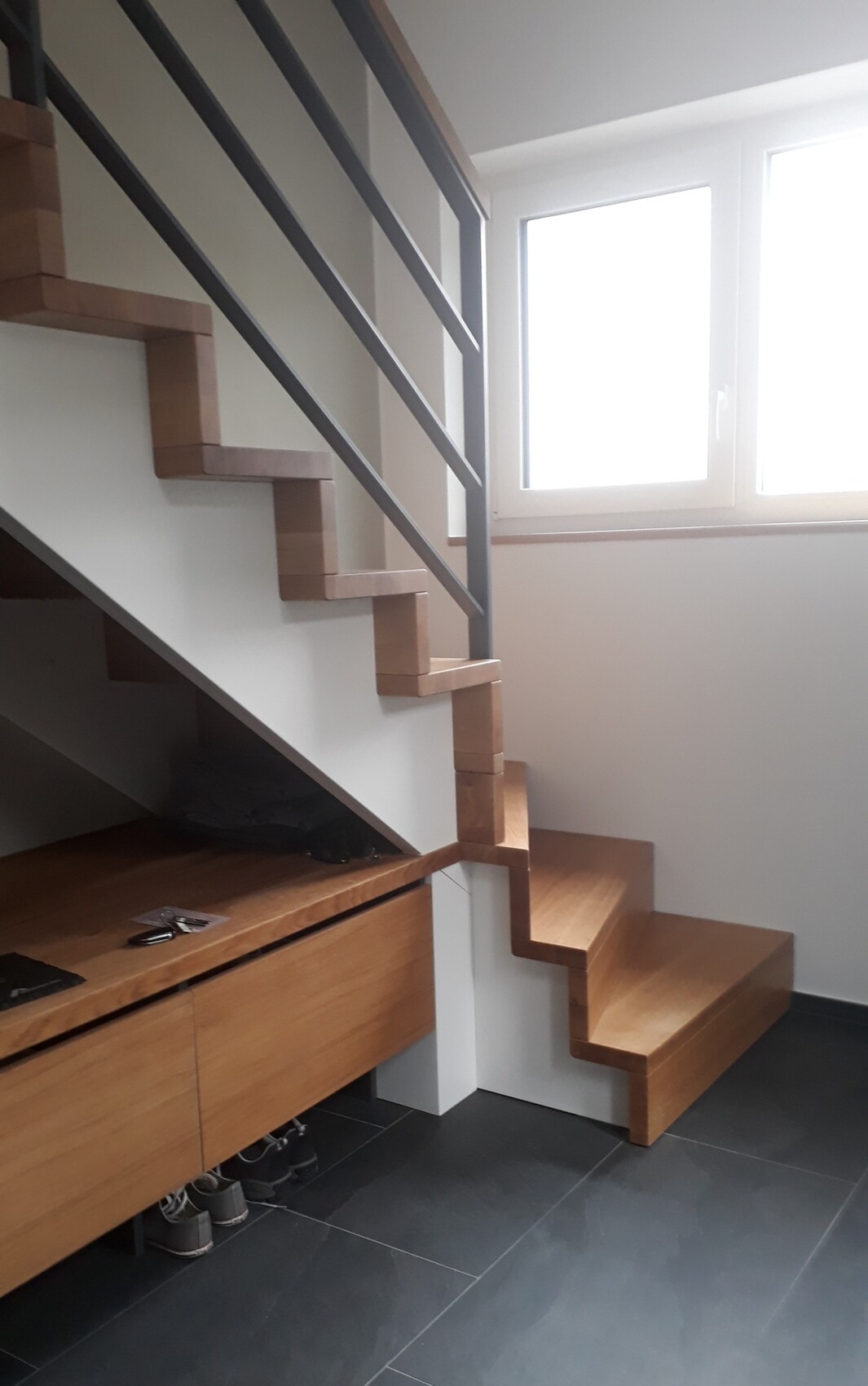 Moderne Innentreppe aus Holzstufen mit schwarzem Geländer, darunter Schränke und Fenster.