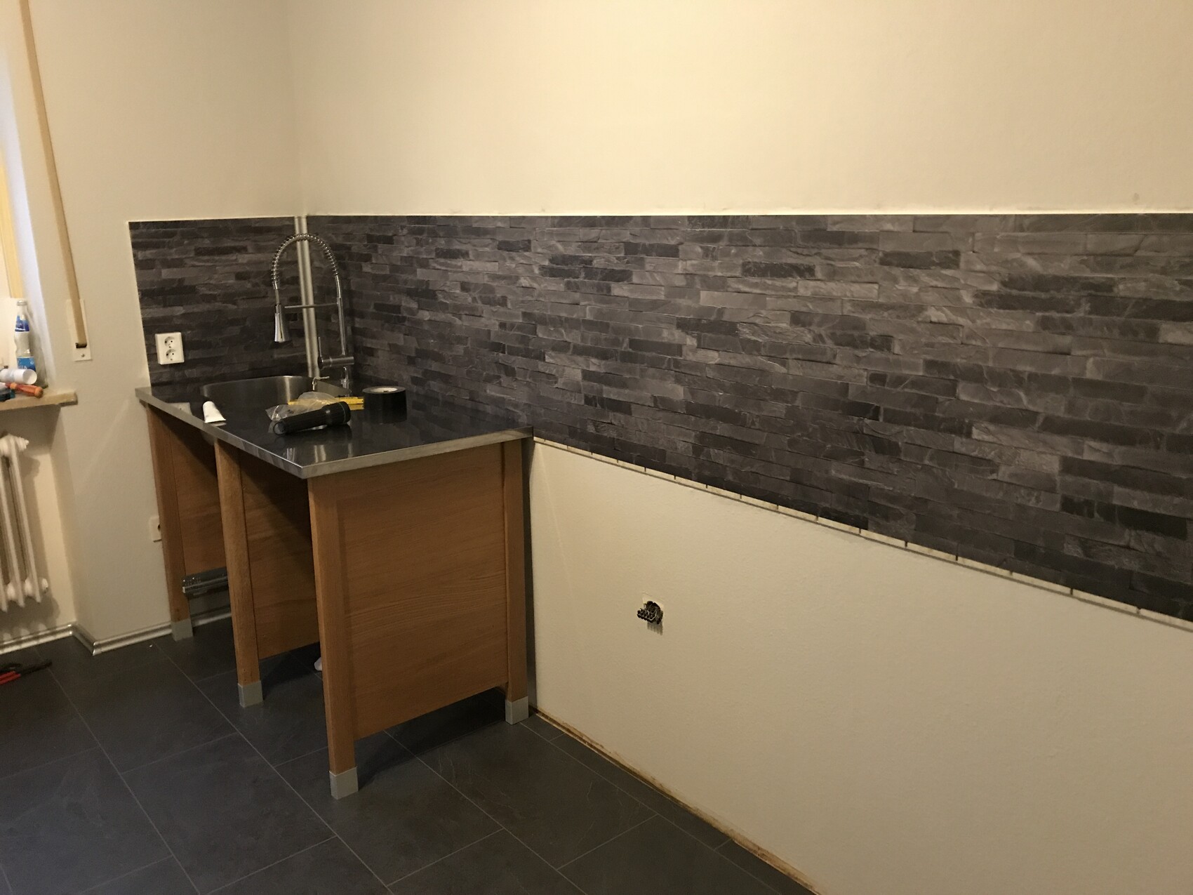 Kitchen with sink and faucet, dark stone backsplash, wooden base cabinet, and gray floor tiles.