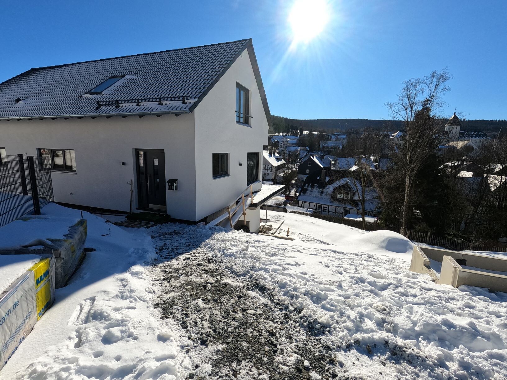 Newly built, white single-family house with dark roof on snow-covered hillside; village in the background.