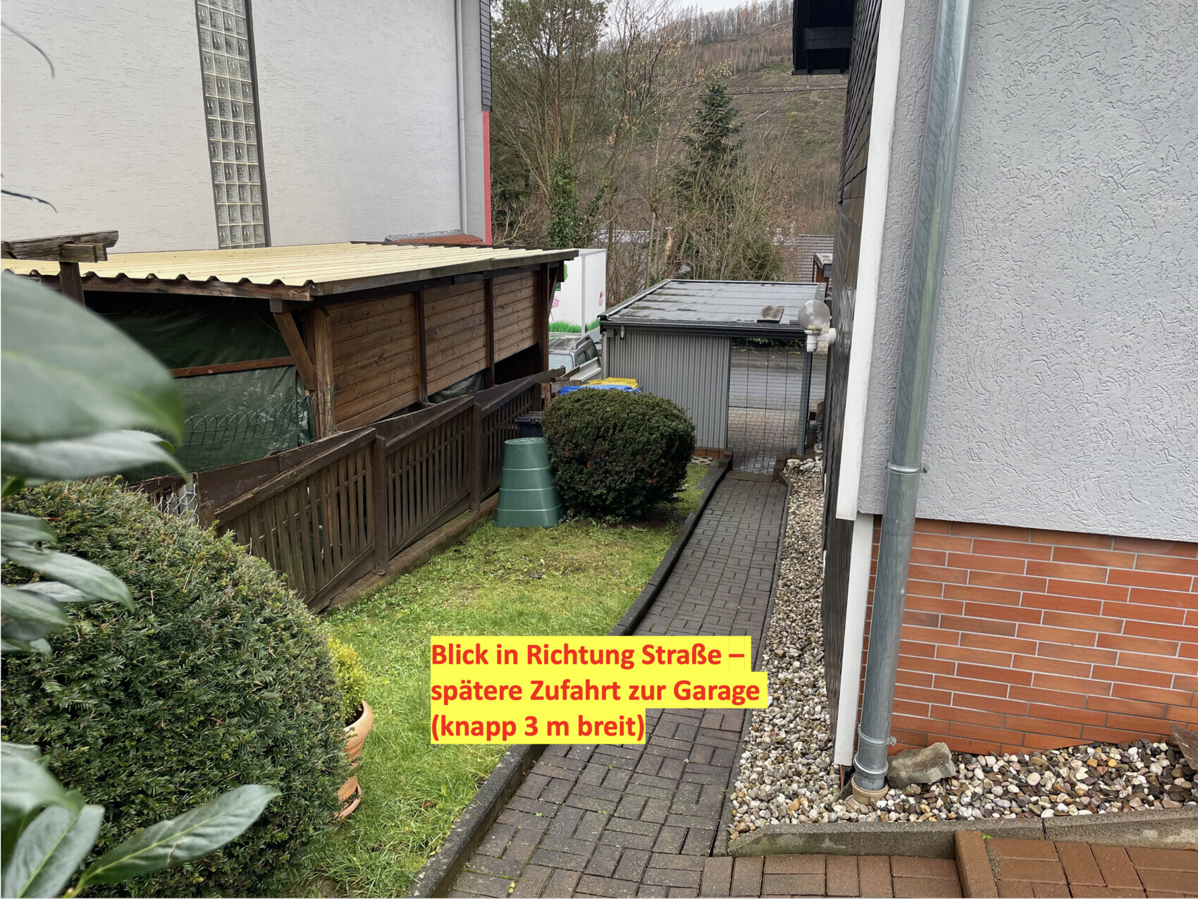 Schmaler Gehweg zwischen Häusern mit Holzzäunen, Busch und Zufahrt zur Garage am Ende