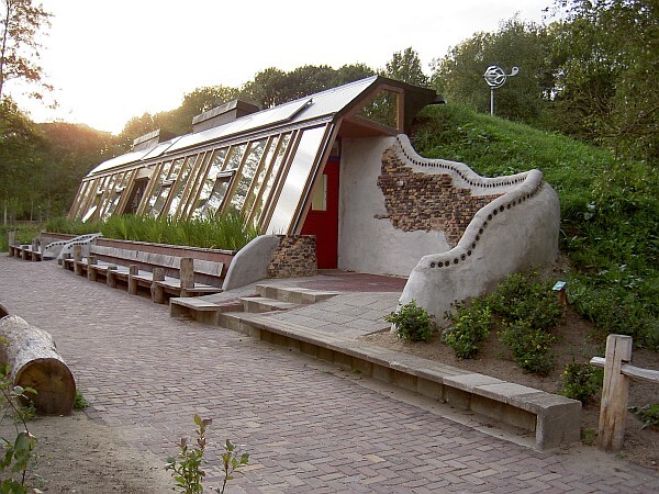 Long greenhouse with sloped roof, next to a paved path alongside a semicircular concrete wall.