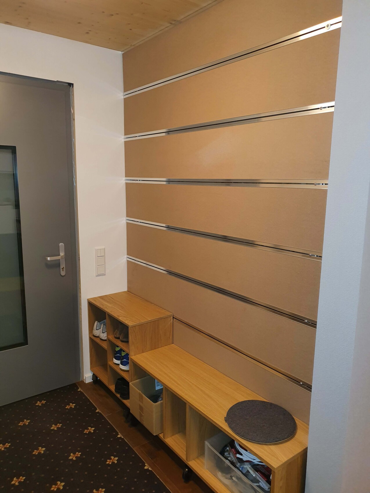 Entrance area with wooden shoe rack, bench, round seat cushion, beige wall panels, and gray door.