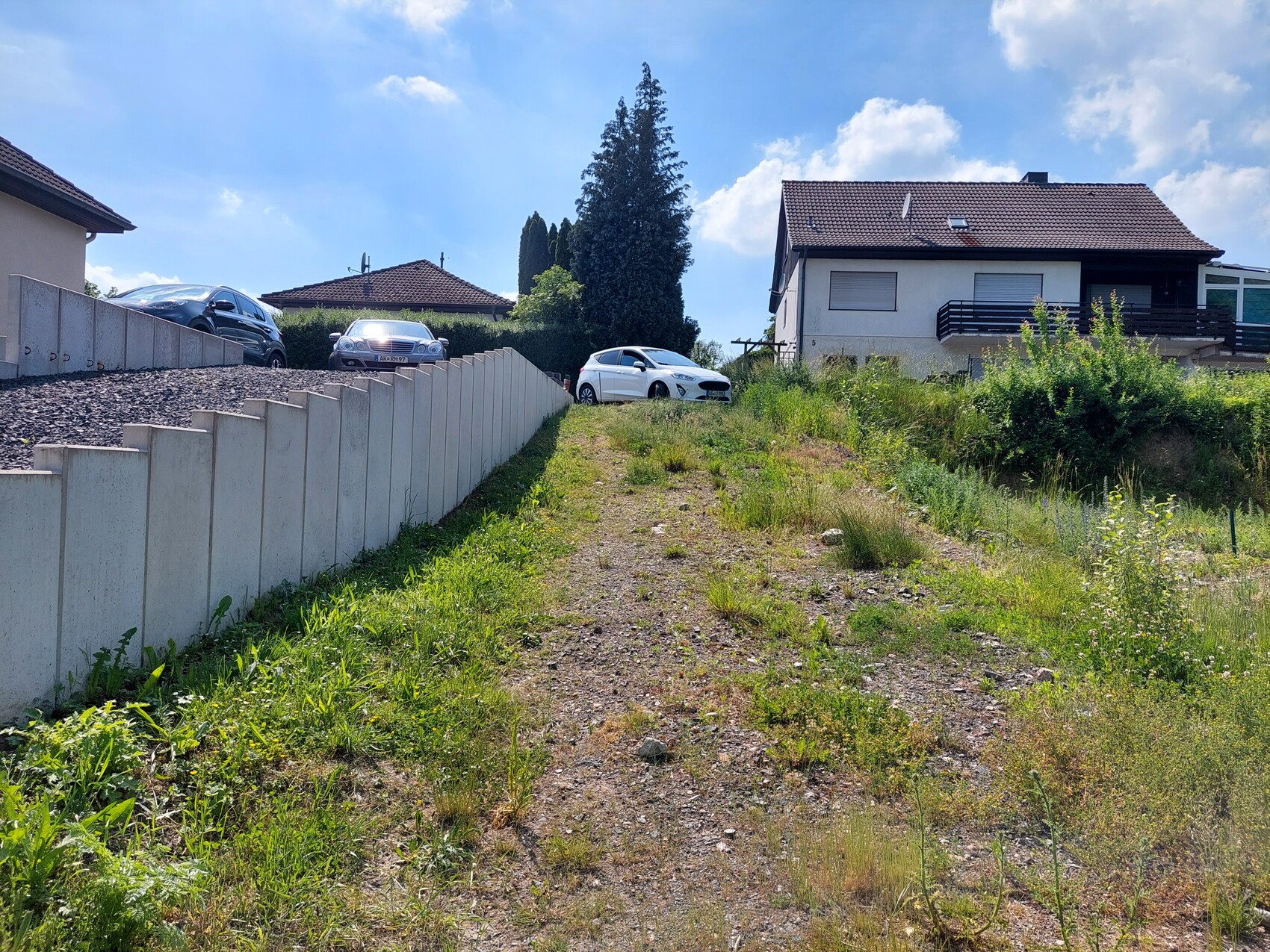 Suburban street with concrete wall on the left, parked cars, and grassy undeveloped land