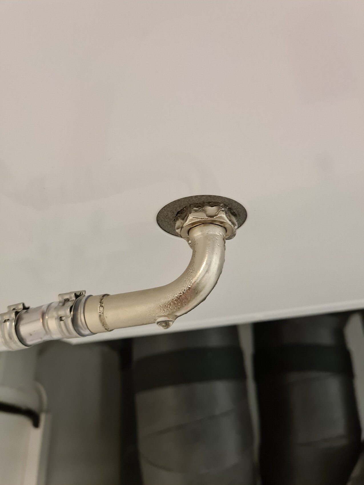 Metal pipe bend with flange on white ceiling surface, with more pipes visible in the background.