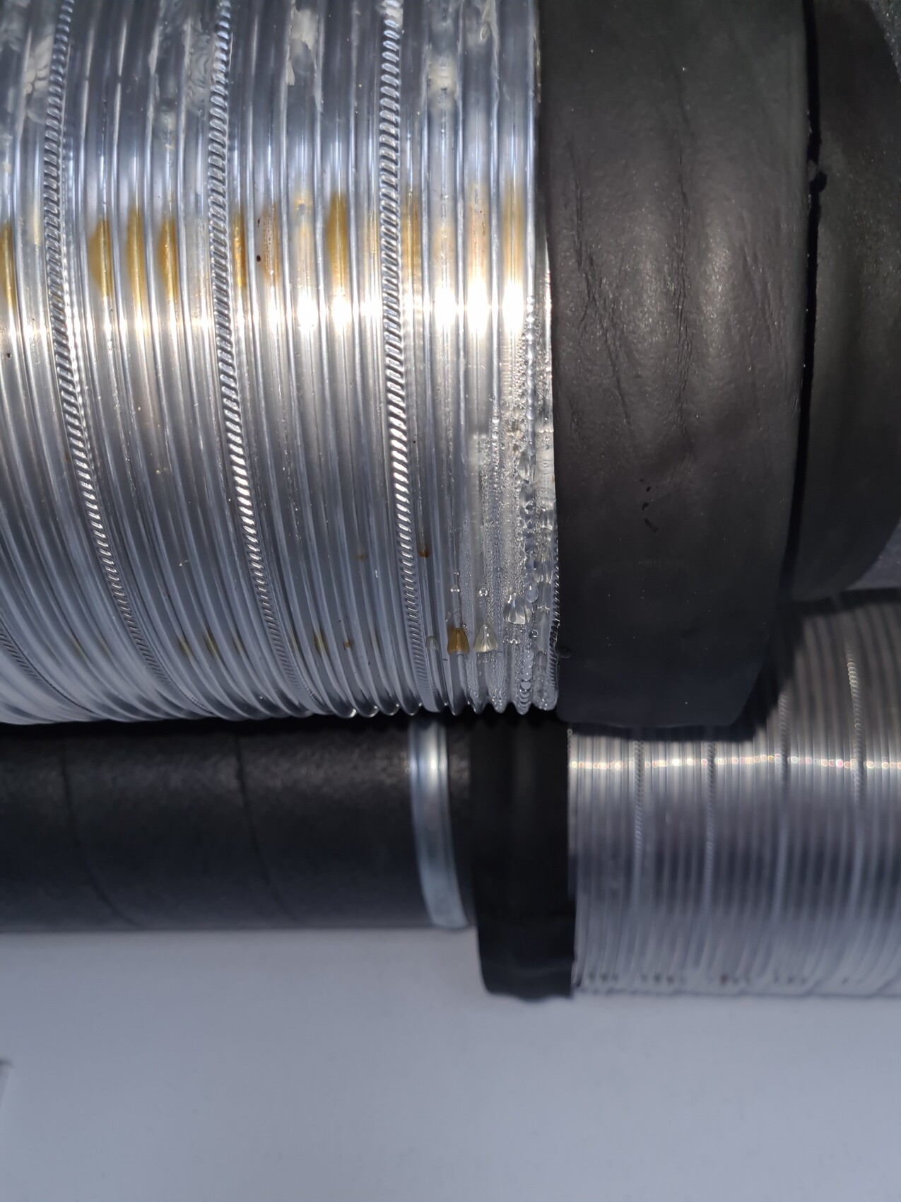 Close-up of a flexible silver metal hose with spiral waves next to a black pipe.