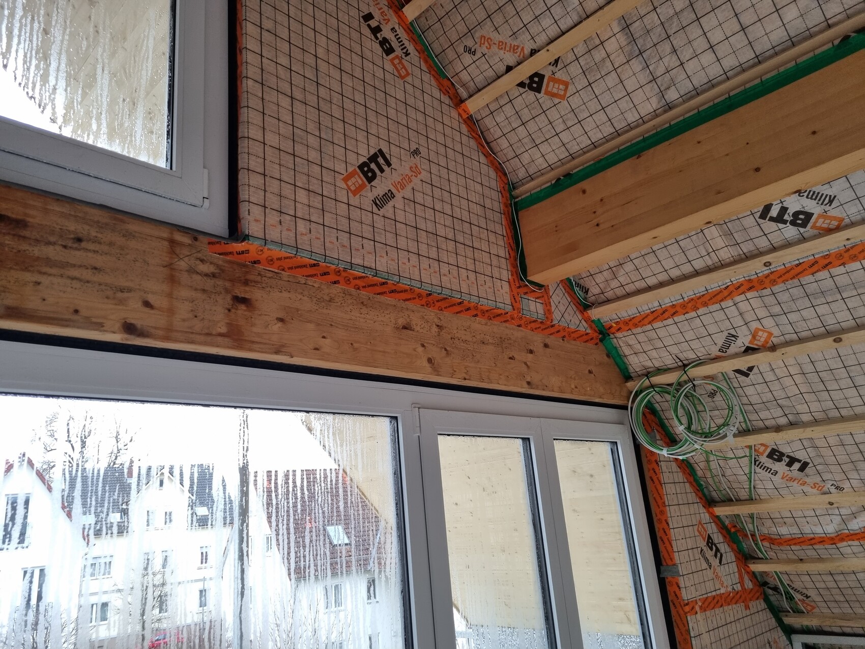 Bau-Dachgeschoss: Dämmplatten, Holzbalken, orange Klebeband, grüne Streifen, Fensterblick auf Häuser