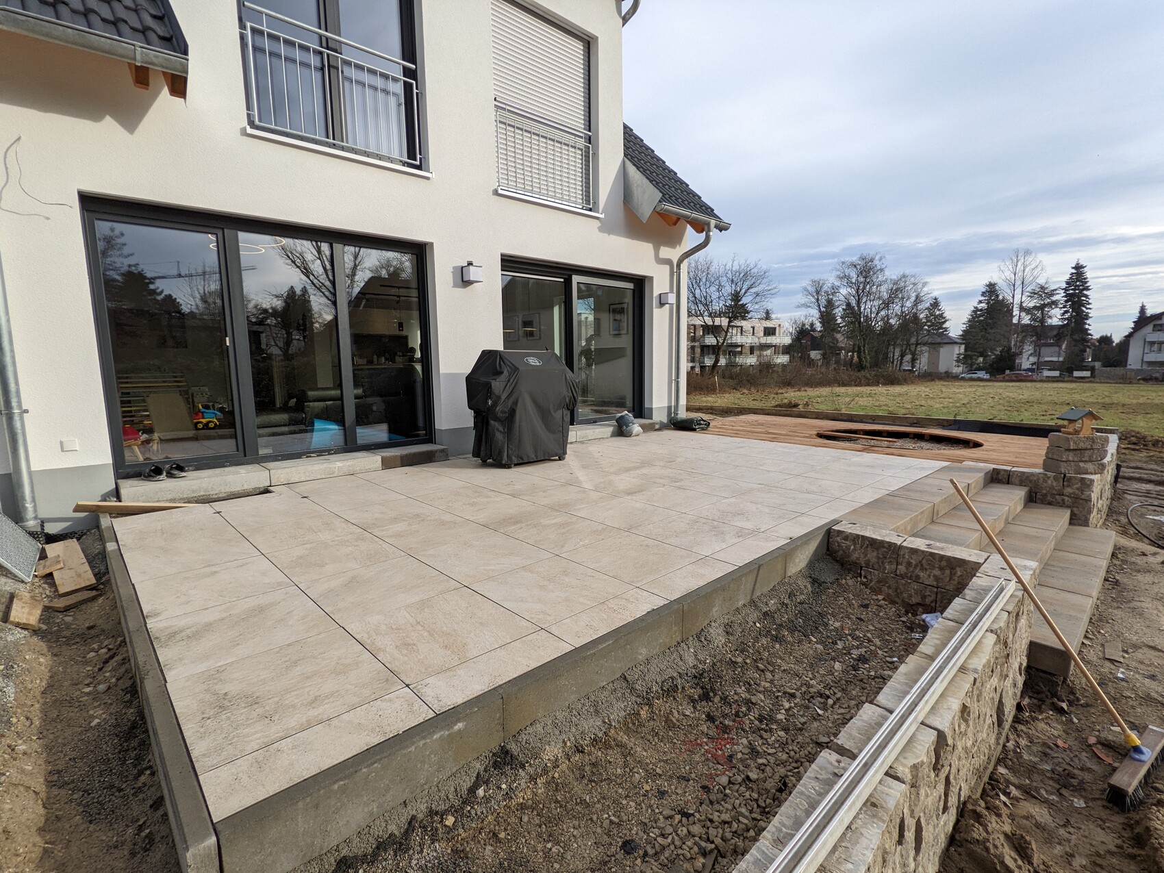 Large tile terrace in front of a modern house; covered grill, construction work in the yard.