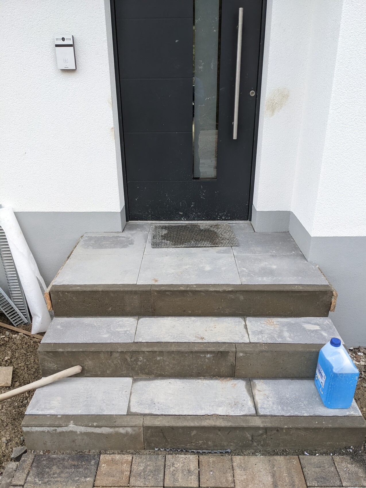 Entrance: black door with glass strip, intercom on the left, steps with mat; blue cleaning bottle on the right.