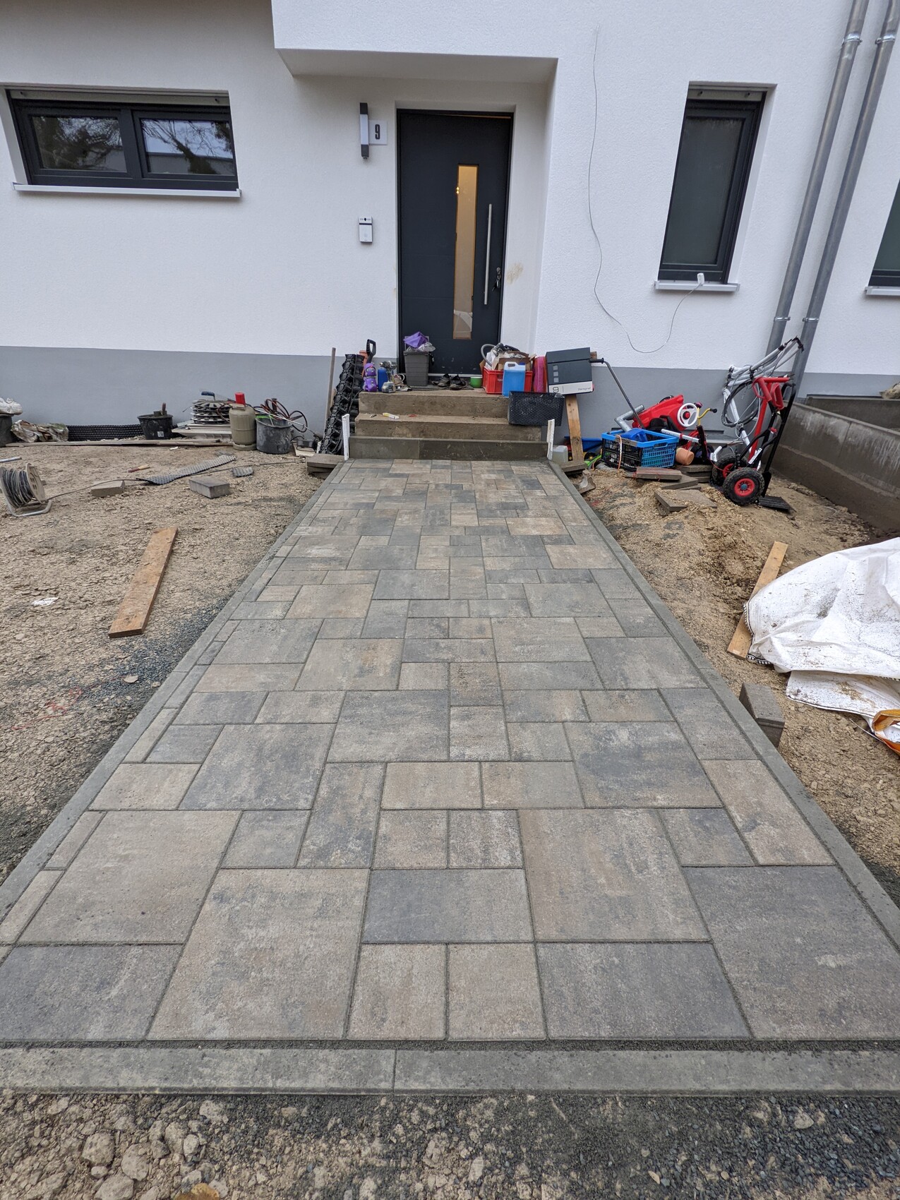 Newly paved walkway made of gray rectangular slabs leads to the front door; construction tools next to it.