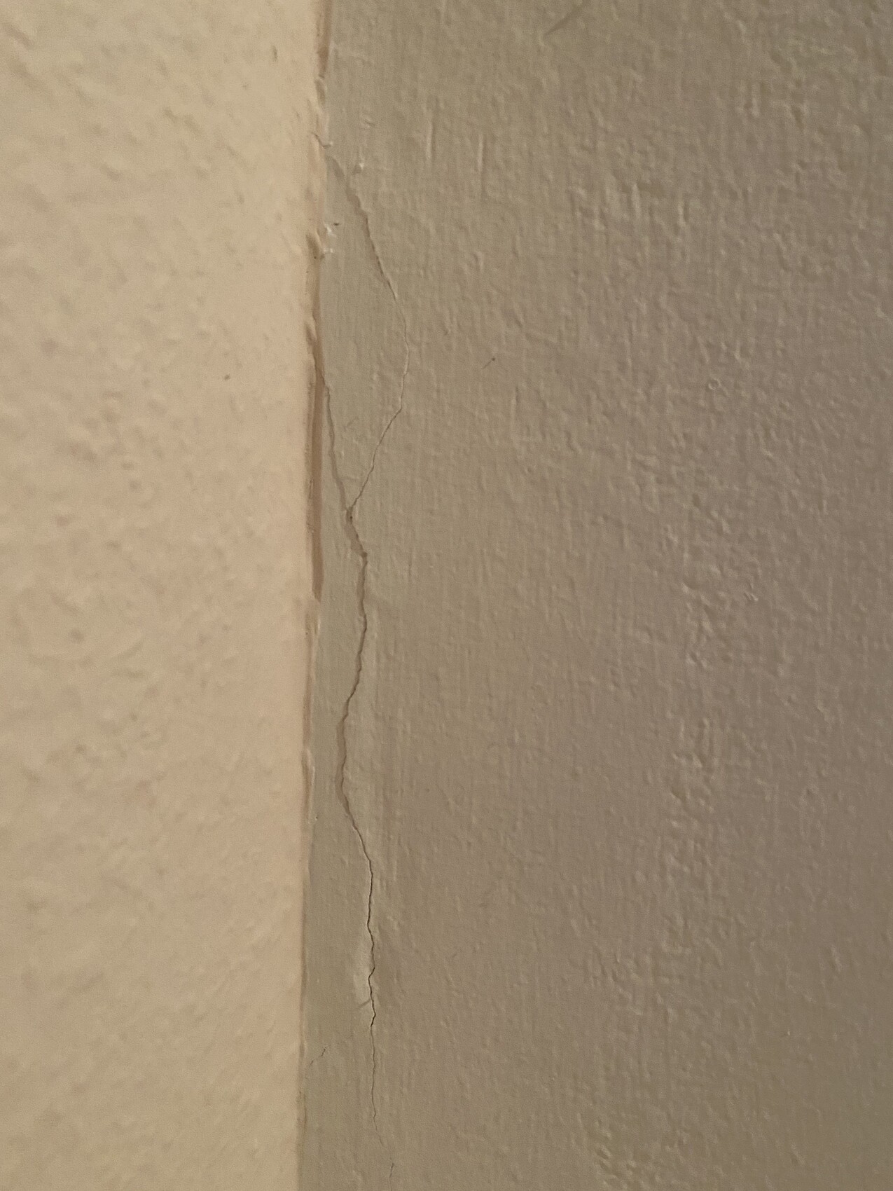 Close-up of a wall corner with a vertical crack between two beige painted surfaces.