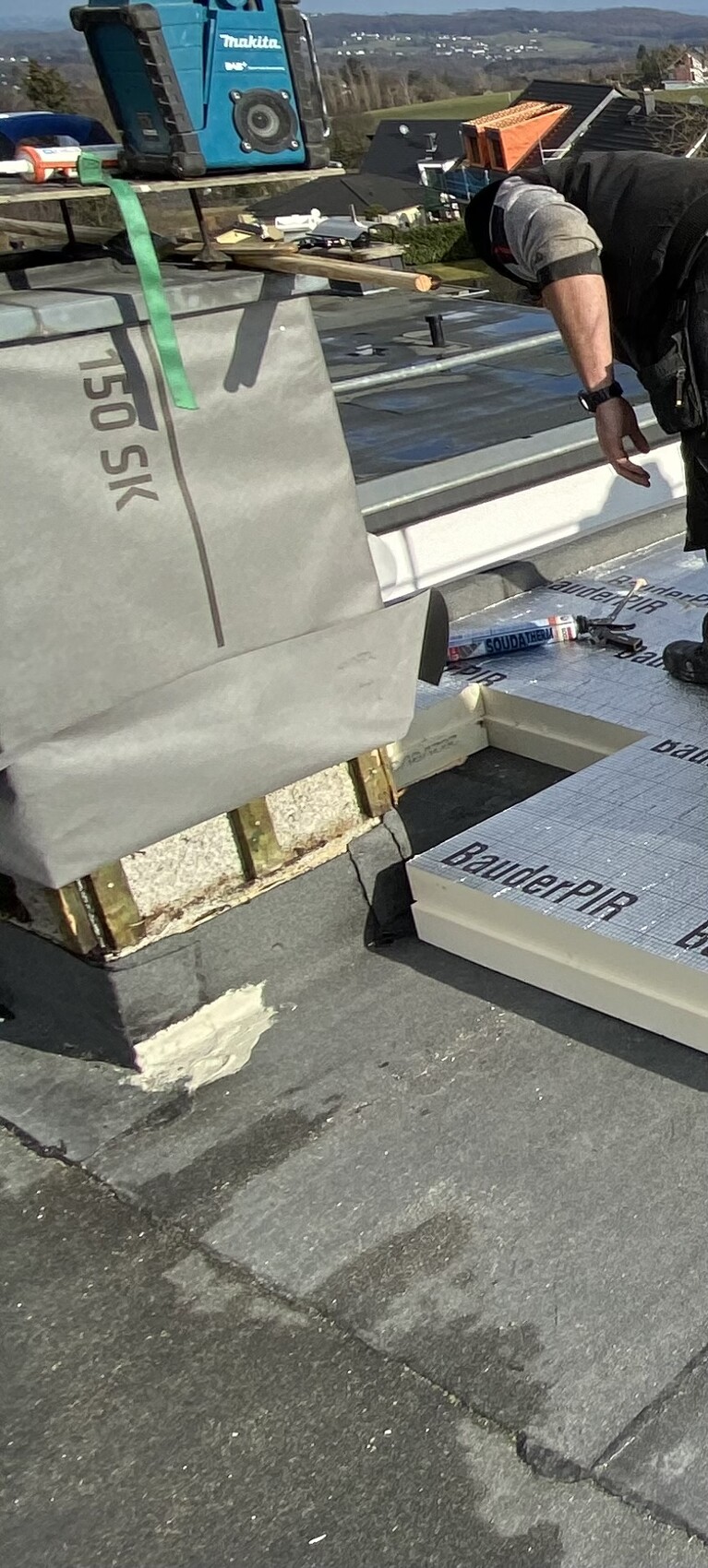 Worker on flat roof installing insulation boards; tools and BauderPIR material visible.
