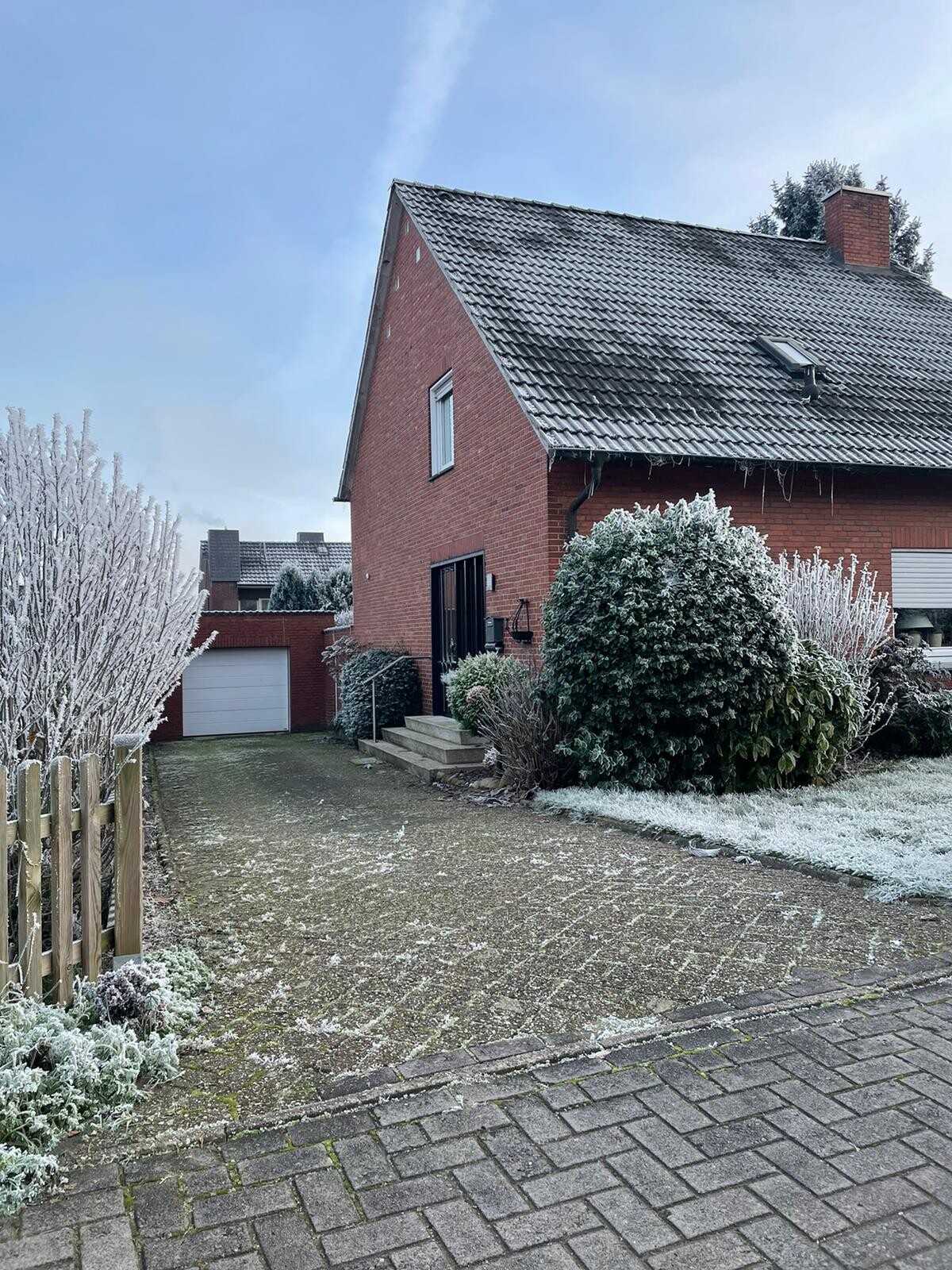 Zweistöckiges rotes Ziegelhaus mit verschneiter Einfahrt, Garage links und frostigen Büschen.