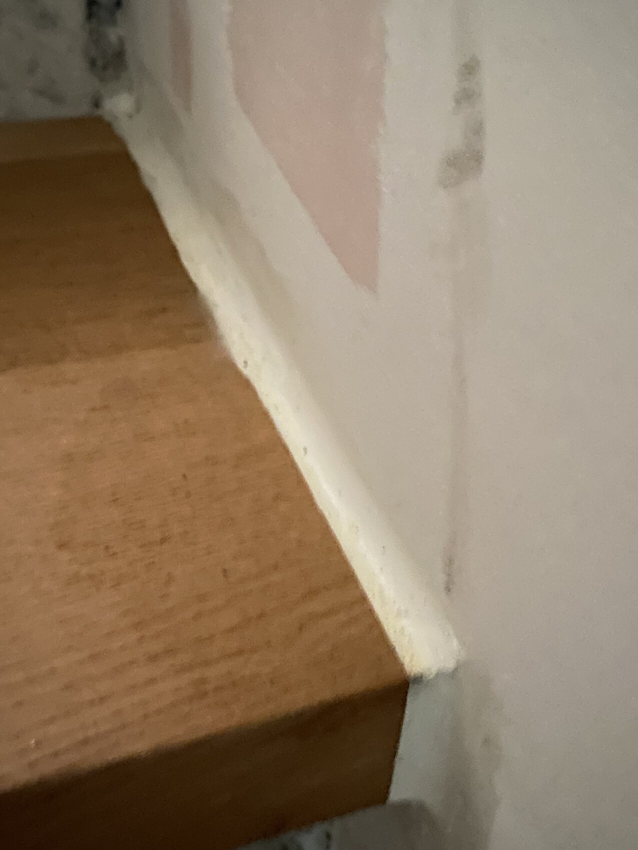 Lower staircase landing made of wood next to a white wall with baseboard; wall with a pink painted area.