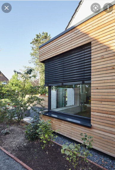 Modern wooden house with large window front, black roller shutters, and garden in front.