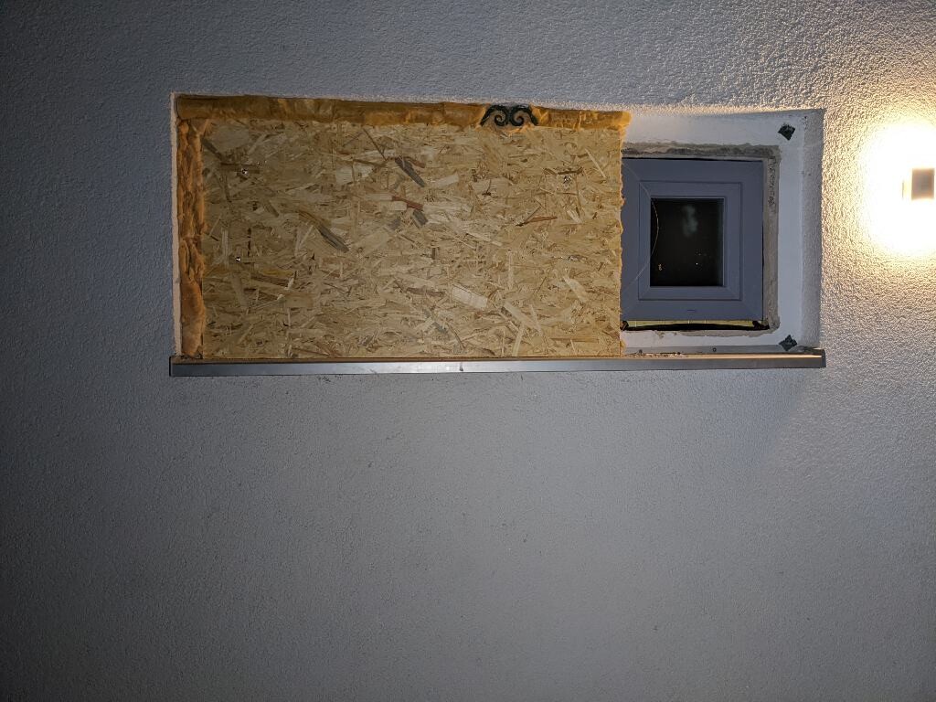 Exterior wall with small blue-framed window; OSB board on the left, outdoor lamp on the right.
